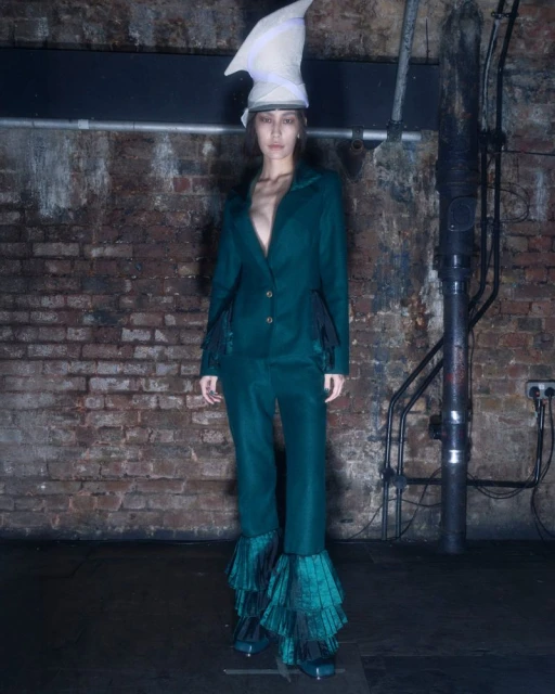 A full shot of a young adult woman in a green suit and unusual hat stands in front of a brick wall with industrial pipes and fixtures.