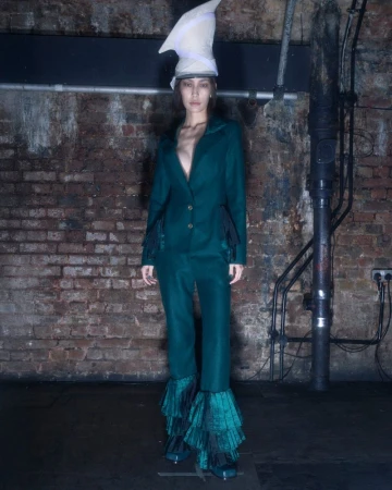 A full shot of a young adult woman in a green suit and unusual hat stands in front of a brick wall with industrial pipes and fixtures.