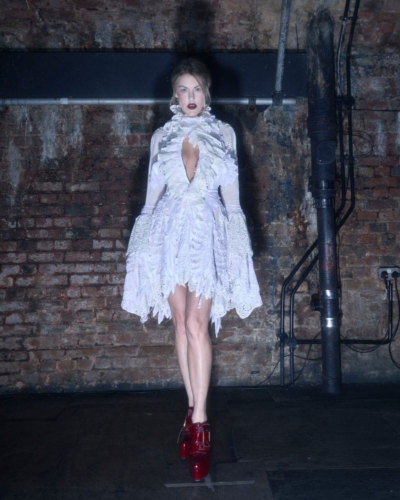 A woman in a frilly white dress and red shoes stands in a dimly lit, industrial-style room with brick walls and dark floors.