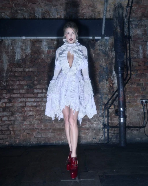 A woman in a frilly white dress and red shoes stands in a dimly lit, industrial-style room with brick walls and dark floors.