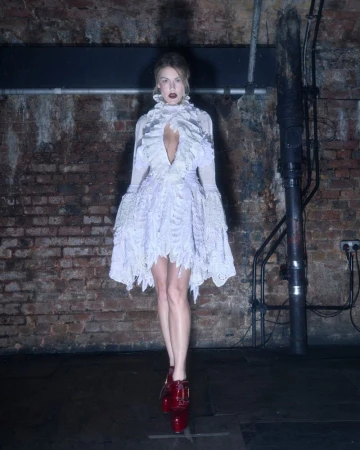 A woman in a frilly white dress and red shoes stands in a dimly lit, industrial-style room with brick walls and dark floors.