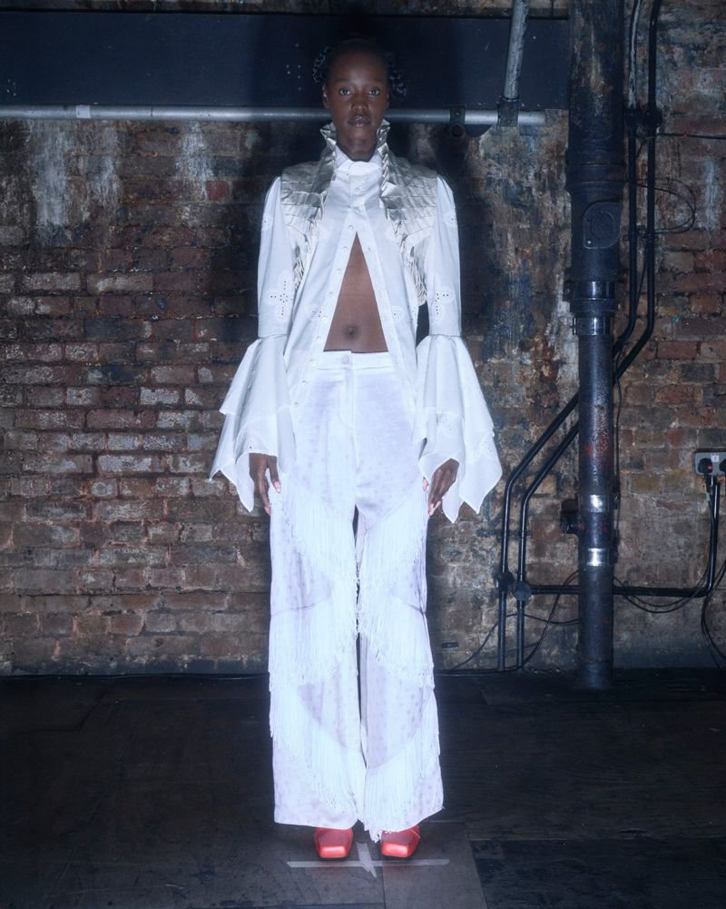 Fashion photo of a Black woman in white bell-sleeved shirt and wide-leg pants against a brick wall, accessorized with statement earrings.