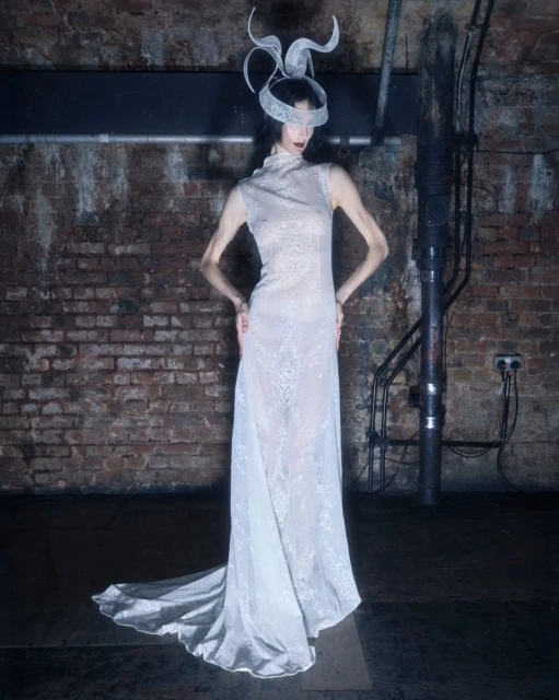 Fashion model in a long white dress and an elaborate white headpiece poses in front of a distressed brick wall indoors.