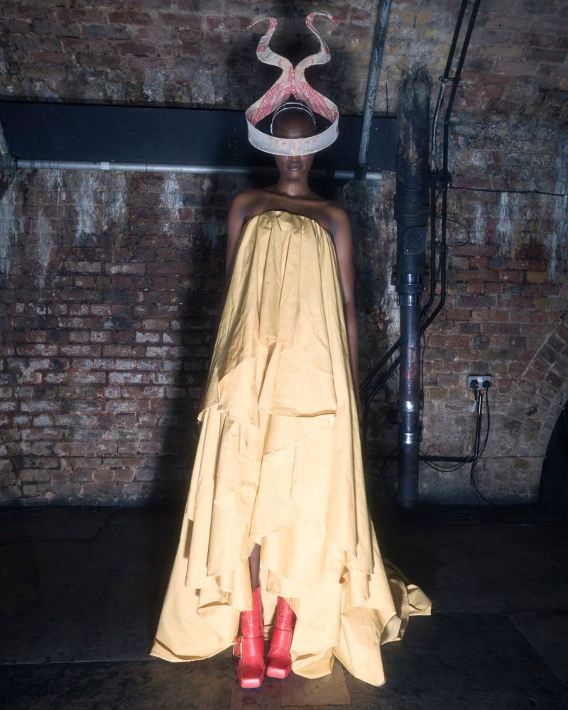 Model wearing a yellow strapless gown and elaborate sculptural headpiece standing against a brick wall.