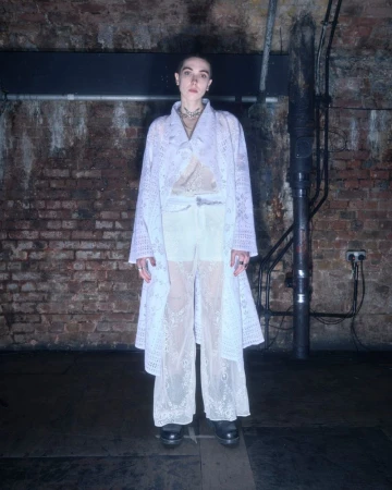 A person with short hair wearing a long white lace coat and matching lace trousers against a dark exposed brick wall.