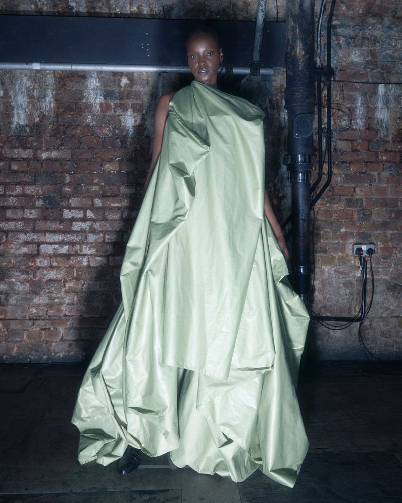 Model wearing a voluminous sage green draped gown against an exposed brick wall interior.