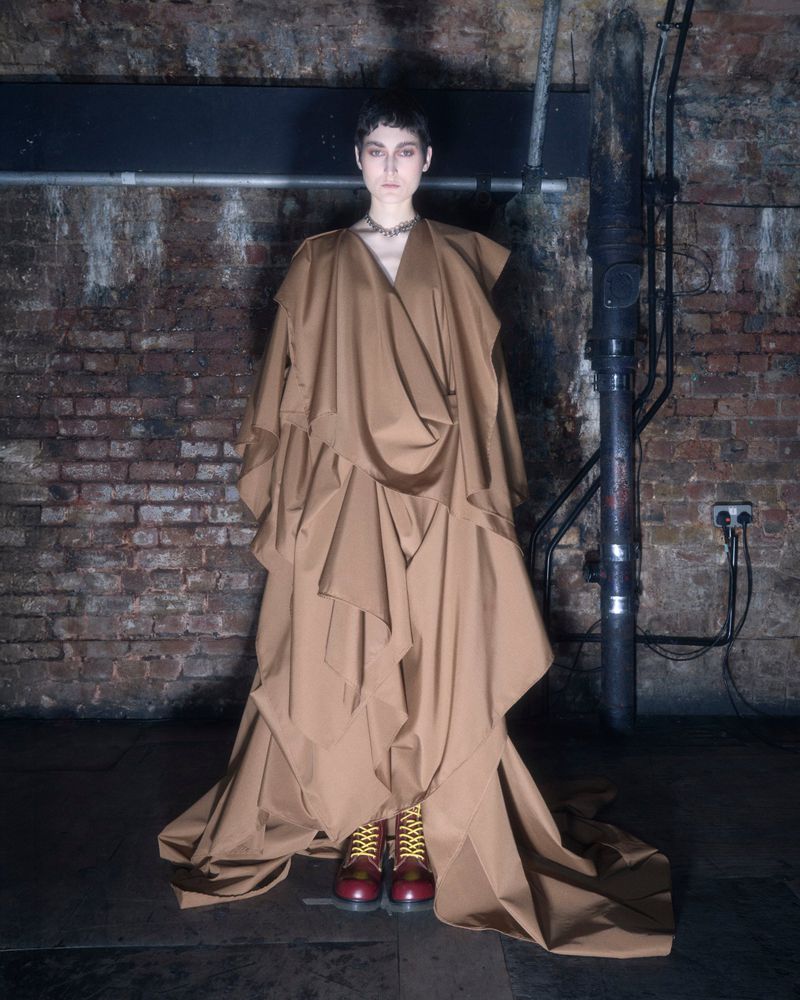 An adult female fashion model stands in a voluminous brown dress and red boots, posed indoors against a brick wall with pipes.