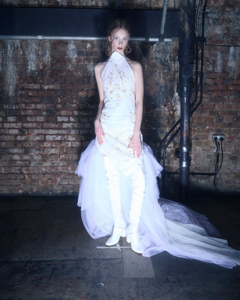 Woman in a white halter dress with a tulle skirt and white thigh-high boots poses in a studio with a brick wall background.