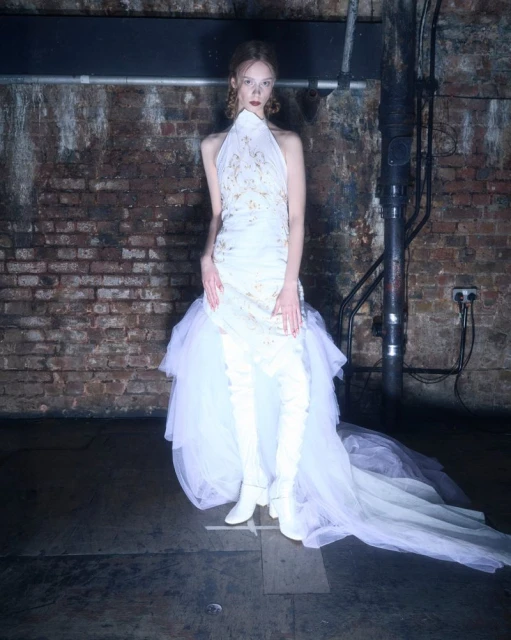 Woman in a white halter dress with a tulle skirt and white thigh-high boots poses in a studio with a brick wall background.