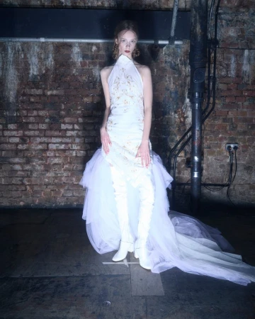 Woman in a white halter dress with a tulle skirt and white thigh-high boots poses in a studio with a brick wall background.