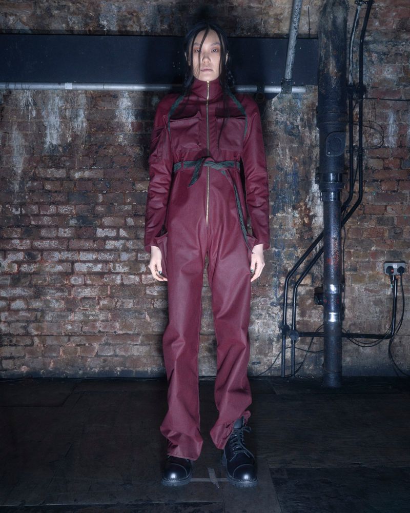 A full shot of an Asian female model wearing a maroon jumpsuit with black boots in front of a brick wall and industrial pipes.