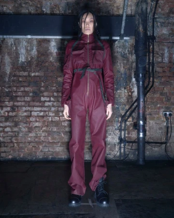 A full shot of an Asian female model wearing a maroon jumpsuit with black boots in front of a brick wall and industrial pipes.