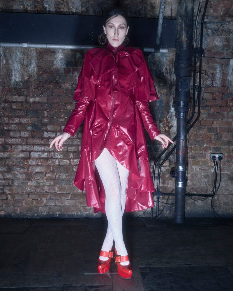 A slender adult woman in a shiny red coat, white tights, and matching platform shoes stands in a gritty urban indoor setting.