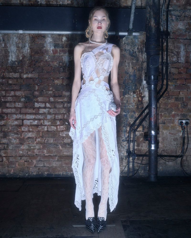 A fair-skinned young adult woman in a white lace dress poses in an indoor setting with dark flooring and a brick wall background.