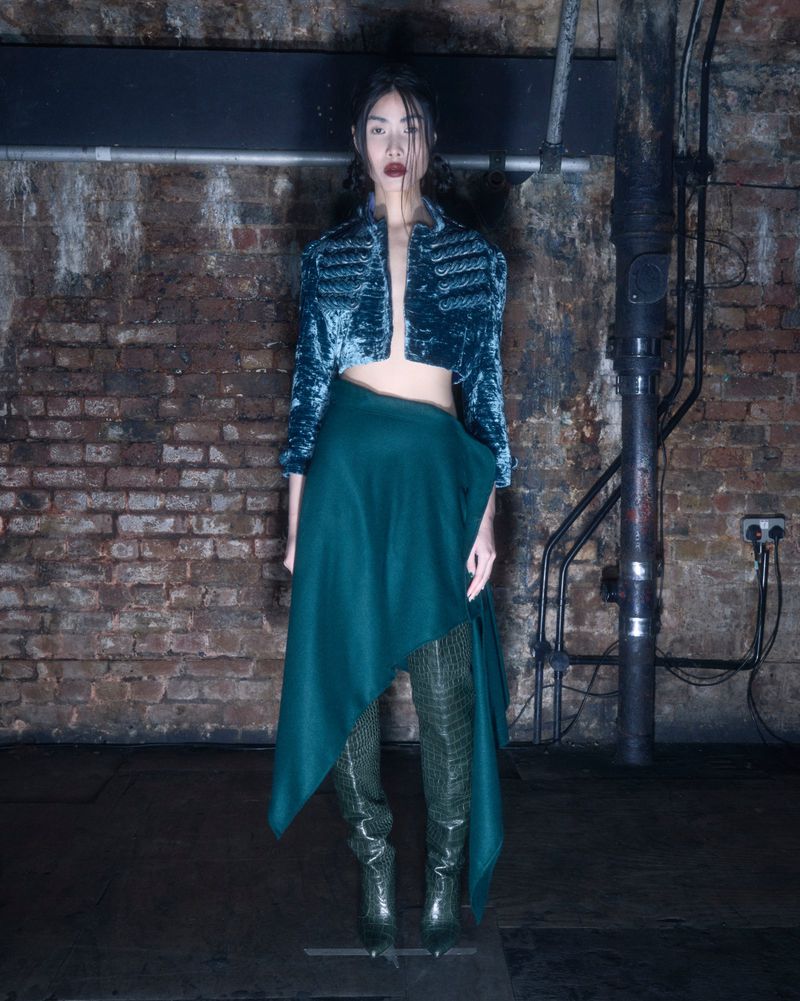 A full-length shot of a slender adult woman in a teal jacket, a green skirt, and green snakeskin boots, posed indoors.