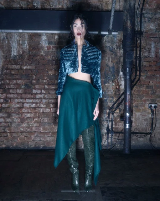 A full-length shot of a slender adult woman in a teal jacket, a green skirt, and green snakeskin boots, posed indoors.