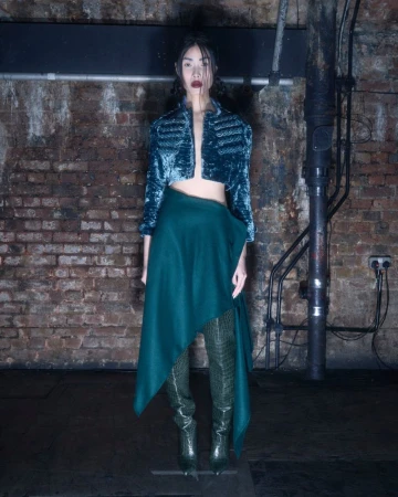 A full-length shot of a slender adult woman in a teal jacket, a green skirt, and green snakeskin boots, posed indoors.