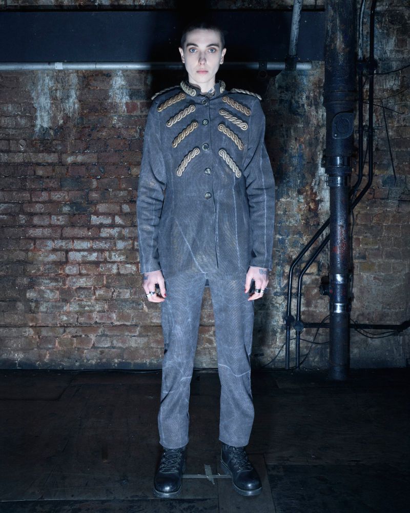 Full-length portrait of a person in a grey military-style jacket and pants, black boots, standing against a weathered brick wall.