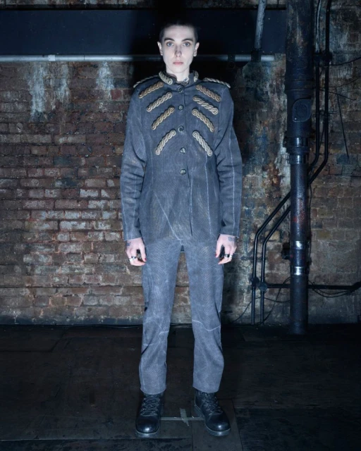 Full-length portrait of a person in a grey military-style jacket and pants, black boots, standing against a weathered brick wall.