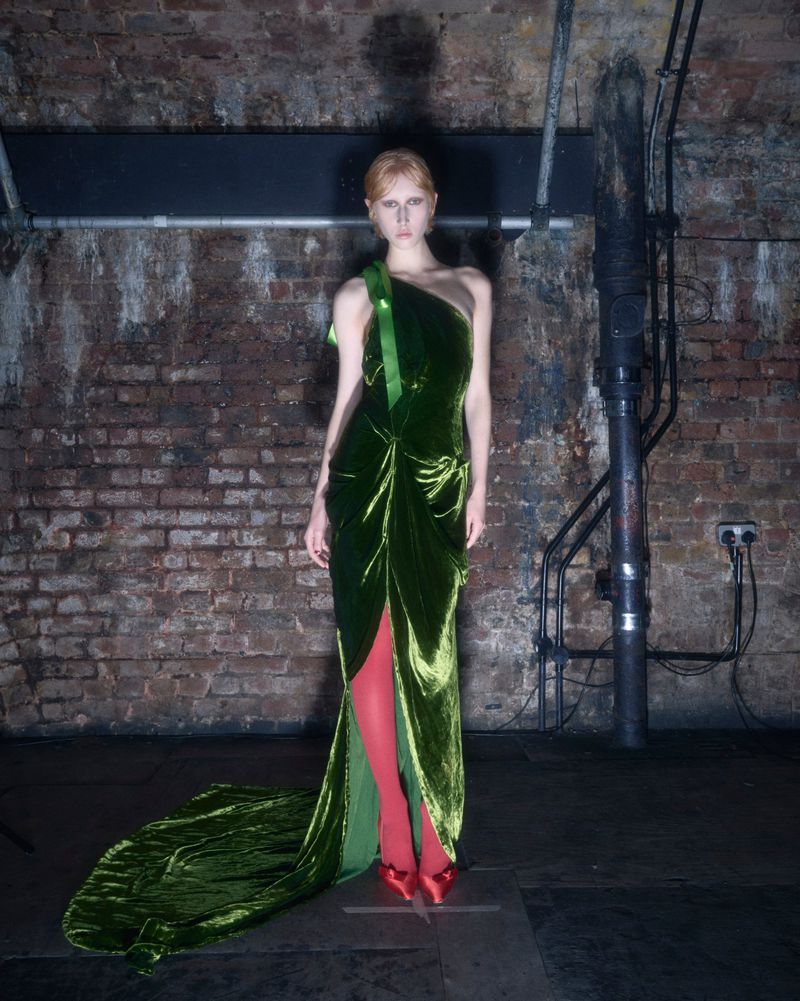 A model in a green velvet gown with red shoes poses against a brick wall with pipes and industrial elements in the background.