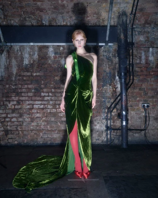 A model in a green velvet gown with red shoes poses against a brick wall with pipes and industrial elements in the background.