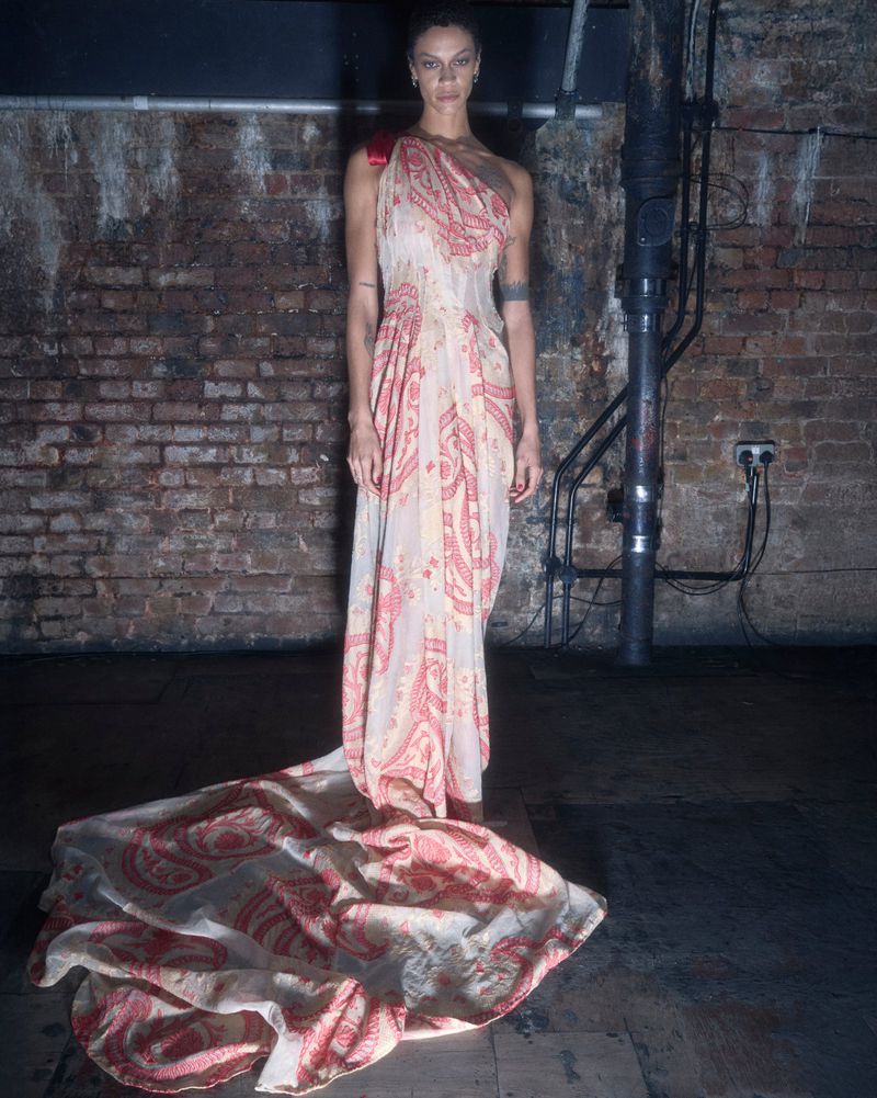 Elegant woman in a patterned gown, standing indoors against a textured brick wall and pipe, creating a timeless, fashionable portrait.