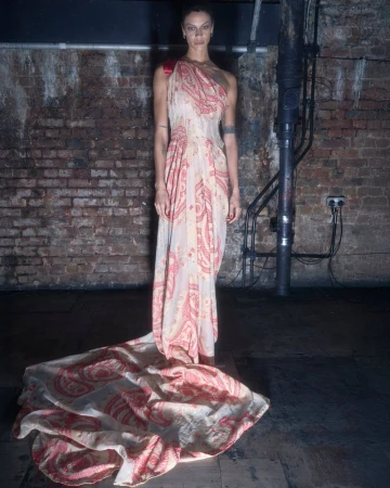 Elegant woman in a patterned gown, standing indoors against a textured brick wall and pipe, creating a timeless, fashionable portrait.
