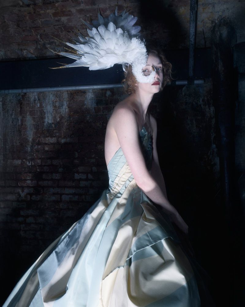 A woman poses in a light blue and tan gown with an elaborate feathered headpiece, set against a dark brick wall, captured in studio lighting.
