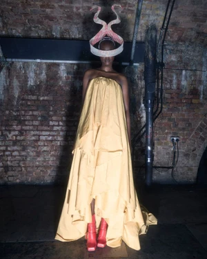 Model wearing a yellow strapless gown and elaborate sculptural headpiece standing against a brick wall.