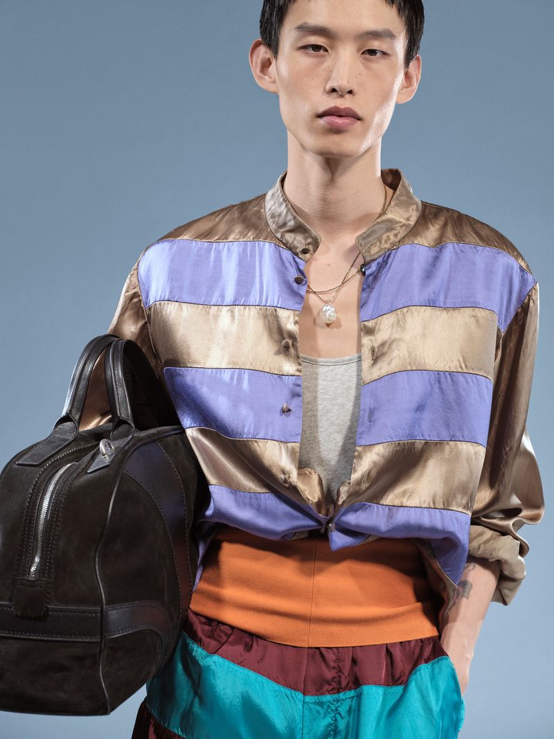 Fashion shot of a stylish adult male wearing a striped shirt, colorful bottoms and a necklace, carrying a leather bag on blue backdrop.