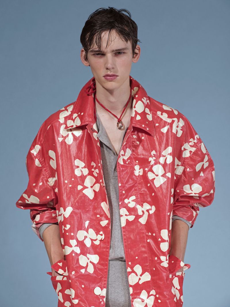 A young adult fashion model poses in a red and white floral-patterned jacket with a grey collared shirt and pendant necklace on a blue background.