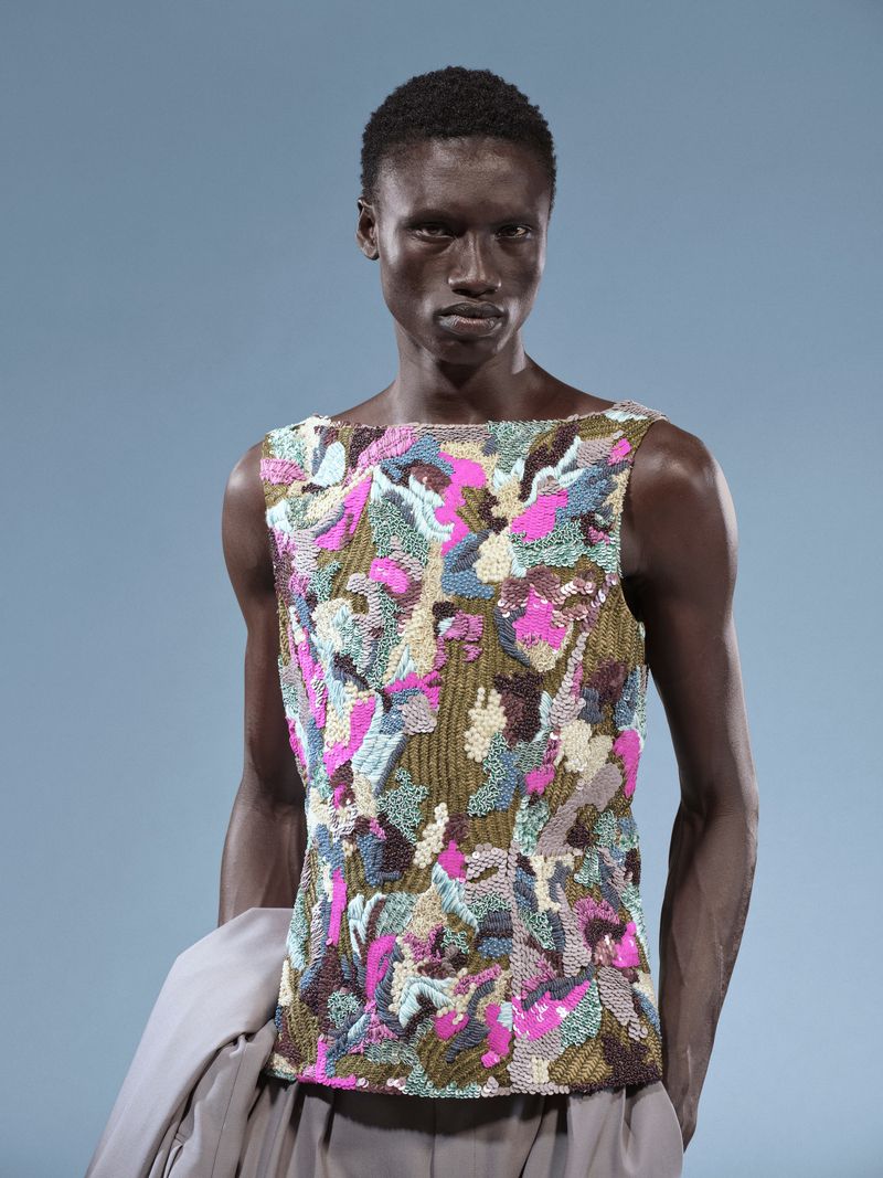 Fashion portrait of a Black male model in a multi-colored embellished sleeveless top against a pale blue studio backdrop, runway fashion.
