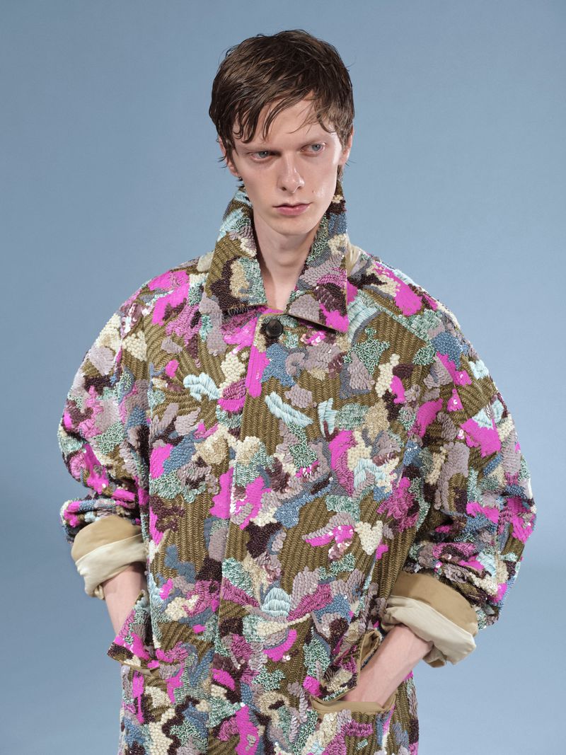 An adult male poses wearing a multicolor print collared jacket with rolled sleeves against a light blue backdrop in a studio setting.
