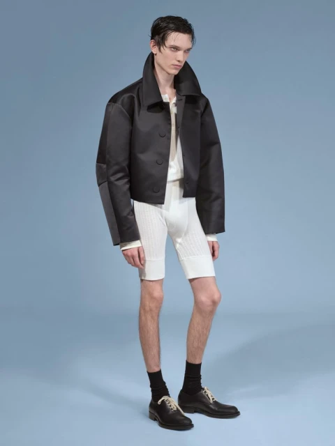 Male fashion model poses in a black jacket and white shorts against a light blue backdrop in a studio fashion shot. 