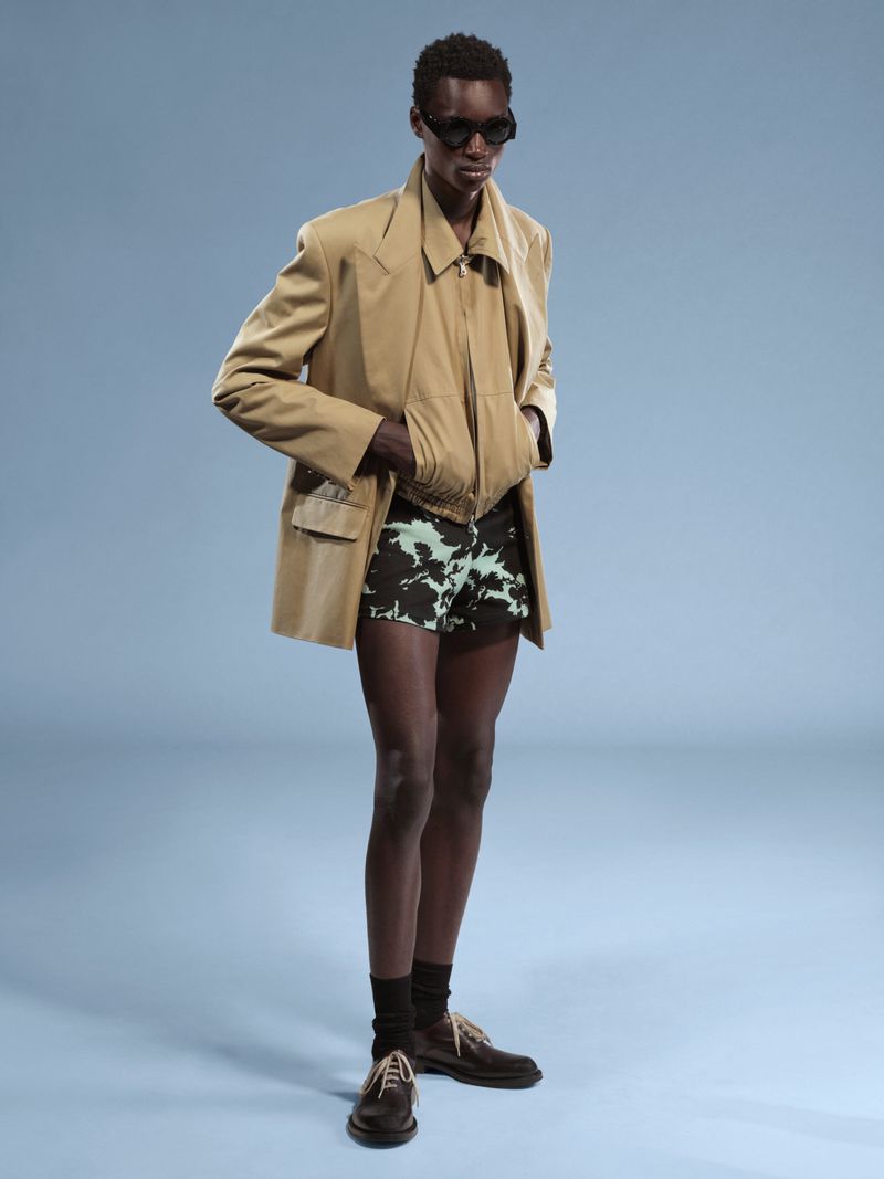 A dark-skinned person in a tan jacket, vest, and patterned shorts poses in front of a light blue background in a full shot.