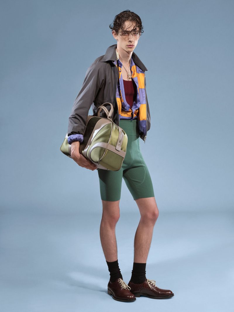 Full-length studio shot of a male fashion model in a gray jacket, patterned top, green shorts, black socks, and brown shoes, carrying a duffel bag.