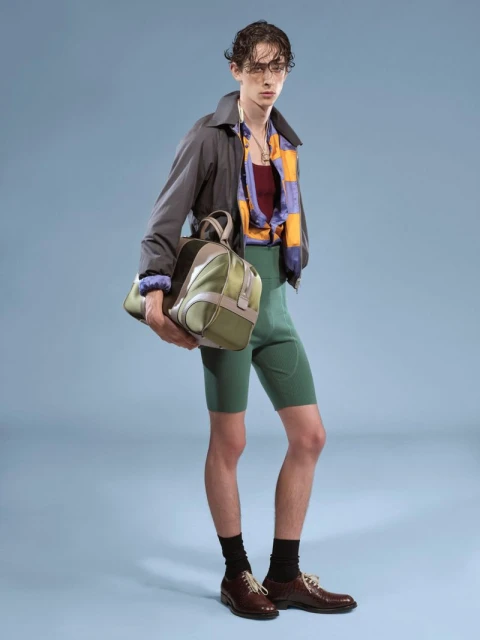 Full-length studio shot of a male fashion model in a gray jacket, patterned top, green shorts, black socks, and brown shoes, carrying a duffel bag.