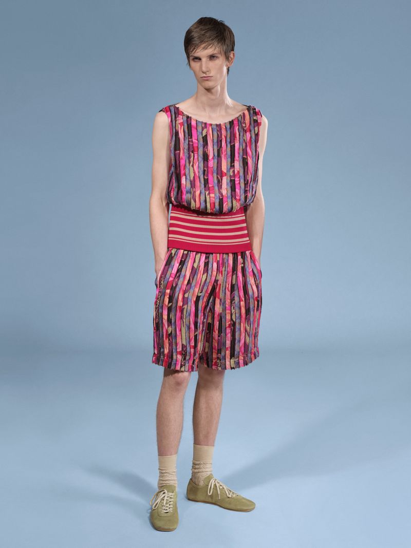 Full shot of a slender person wearing a striped pink and black sleeveless top and matching shorts, posing on a blue backdrop.