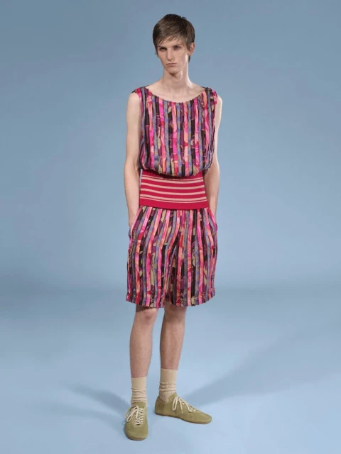 Full shot of a slender person wearing a striped pink and black sleeveless top and matching shorts, posing on a blue backdrop.