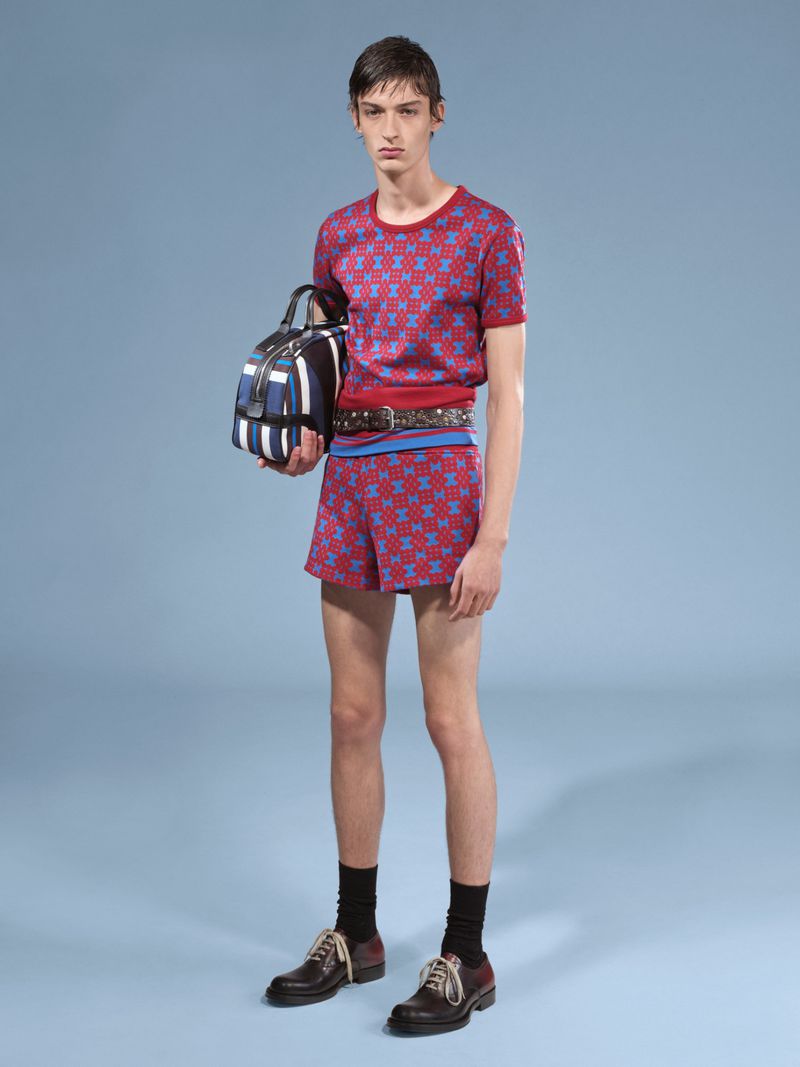 Male fashion model poses in matching red and blue patterned knitwear shorts and t-shirt, holding a striped duffel bag.