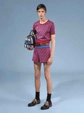 Male fashion model poses in matching red and blue patterned knitwear shorts and t-shirt, holding a striped duffel bag.