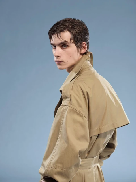 Close-up studio portrait of a male fashion model with dark hair looking over his shoulder, wearing a beige trench coat against a blue background.