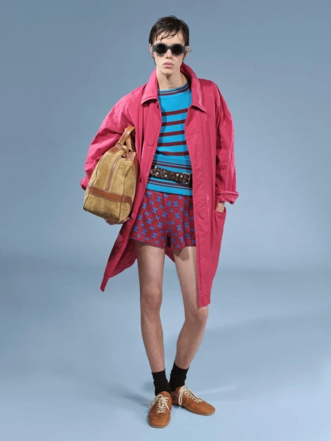 Full shot of a male model in a pink coat and shorts, wearing sunglasses and carrying a duffel bag, on a light blue background.