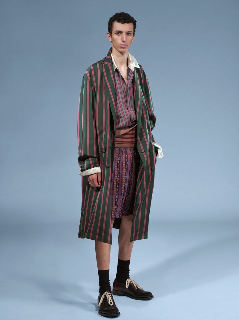 Fashionable adult male model posing in a striped brown and green robe, button-up shirt, and matching shorts on a studio backdrop.