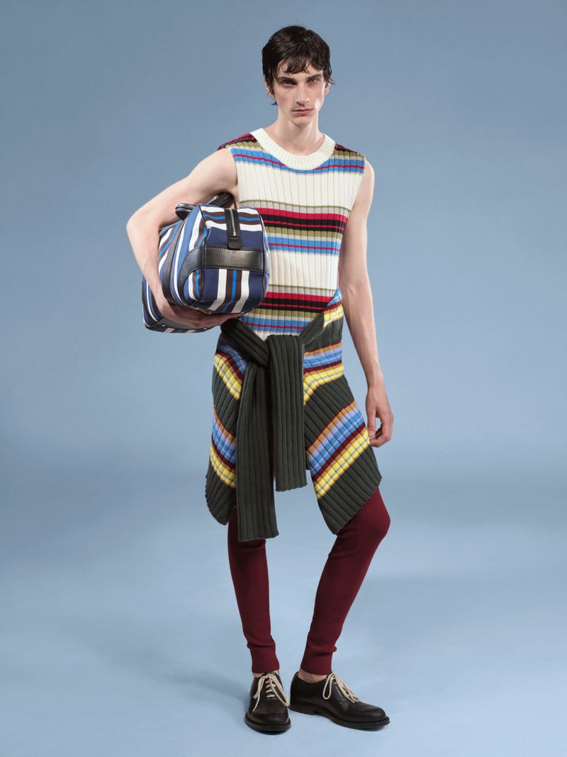 A slender adult male poses in a studio, wearing a striped sleeveless top, dark leggings, black shoes, and carrying a striped duffel bag.