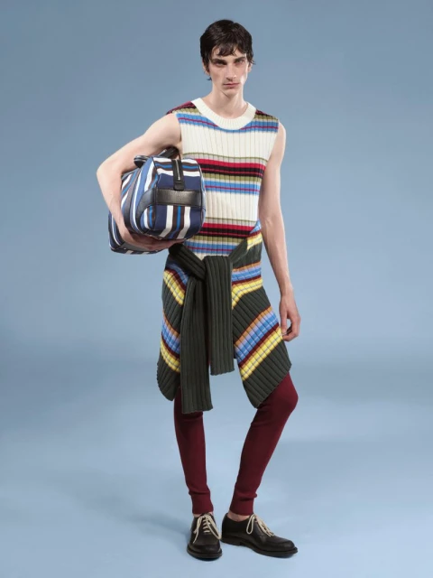 A slender adult male poses in a studio, wearing a striped sleeveless top, dark leggings, black shoes, and carrying a striped duffel bag.
