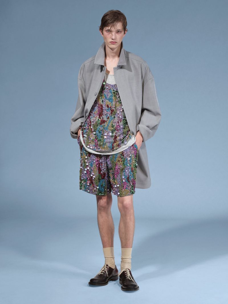 A young adult male stands indoors against a light blue backdrop, wearing a patterned top and shorts with a grey coat and brown shoes.