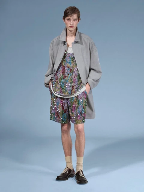 A young adult male stands indoors against a light blue backdrop, wearing a patterned top and shorts with a grey coat and brown shoes.