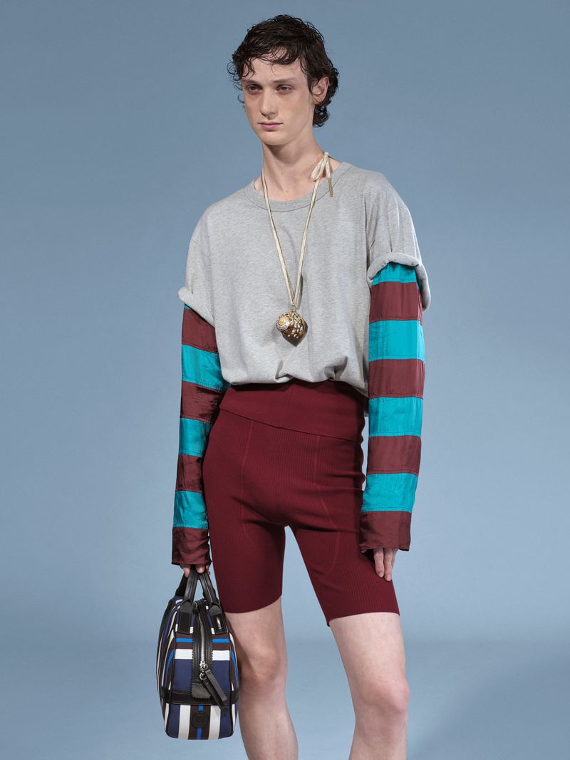 Fashionable male model poses in shorts, striped long sleeve, and grey top against a light blue studio backdrop, holding a striped bag.