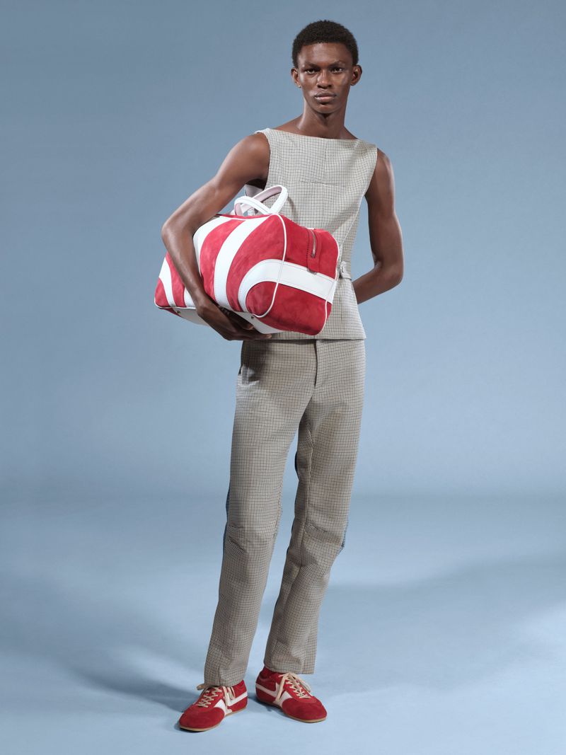Adult male fashion model posing with a striped duffel bag. He is wearing a sleeveless top and pants, paired with matching red and white sneakers.
